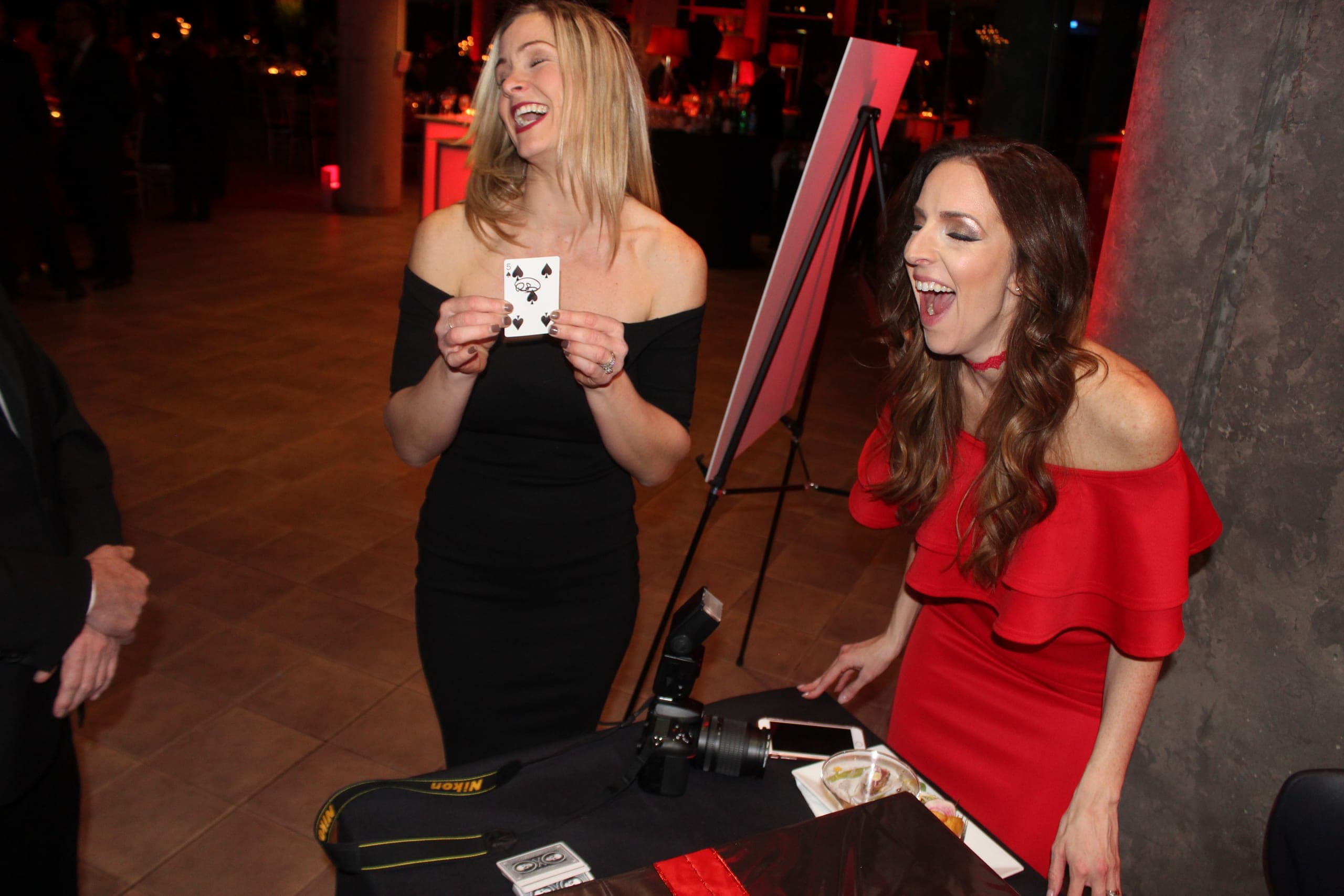 Two guests laughing in amazement as one holds up a signed five of spades at a gala