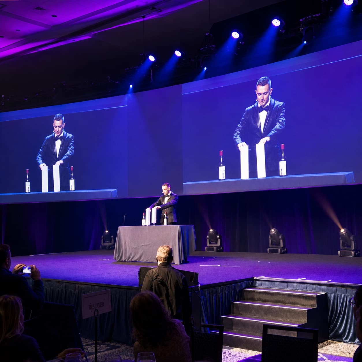 Chris Dare on stage in a tuxedo before a corporate ballroom audience, shown on two large screens
