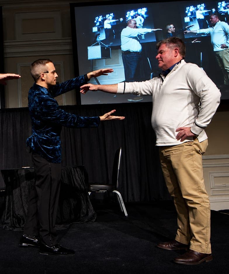 Two volunteers point at each other on stage beside a chained prediction box as Chris Dare directs, shown on twin screens