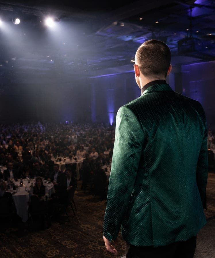 Chris Dare in a green sequined jacket facing a packed ballroom of hundreds from the stage